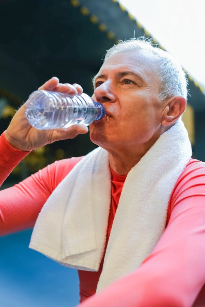 Elderly man wearing red long sleeves and towel, drinking bottled water post-exercise.